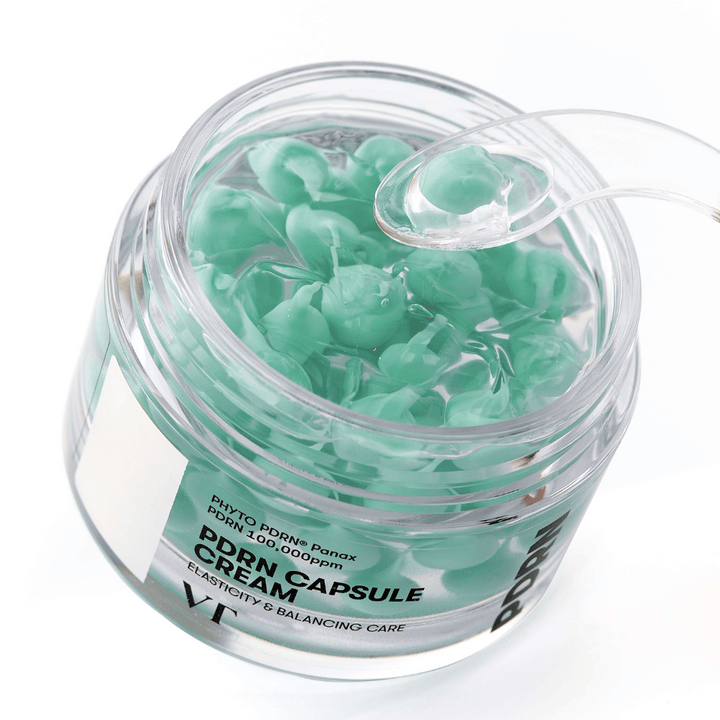 Gros plan sur un pot ouvert de VT PDRN Capsule Cream. Le contenu se compose de petites capsules vertes dans un gel transparent, avec une spatule fournie pour l'application. Le texte sur le pot indique "Elasticity & Balancing Care".