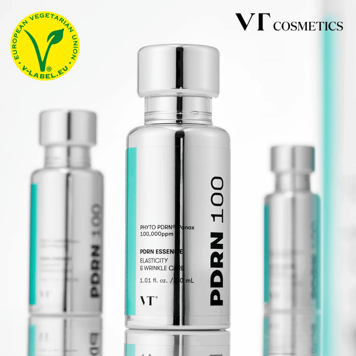 Plusieurs bouteilles argentées de VT PDRN 100 Essence sont alignées, avec un marquage clair de 100,000 ppm PDRN et un accent sur l'élasticité et le soin des rides. En haut, on voit le logo de VT Cosmetics et un label V pour le contenu végétarien.