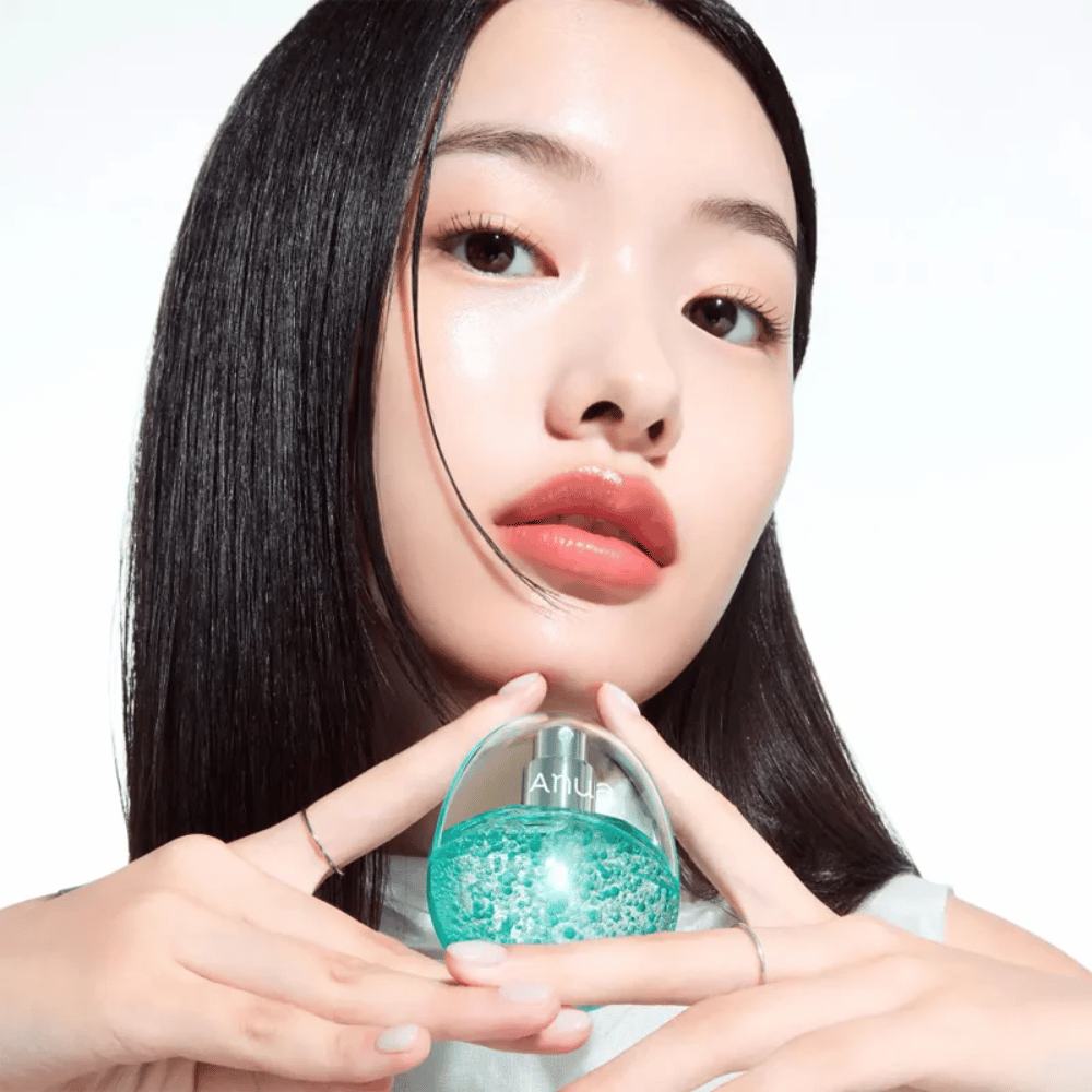 Gros plan sur un modèle tenant un flacon rond et transparent de Anua rempli de capsules ou de perles de gel turquoise. L’image met l’accent sur l’éclat de la peau et le design innovant du produit, donnant une impression fraîche et moderne.