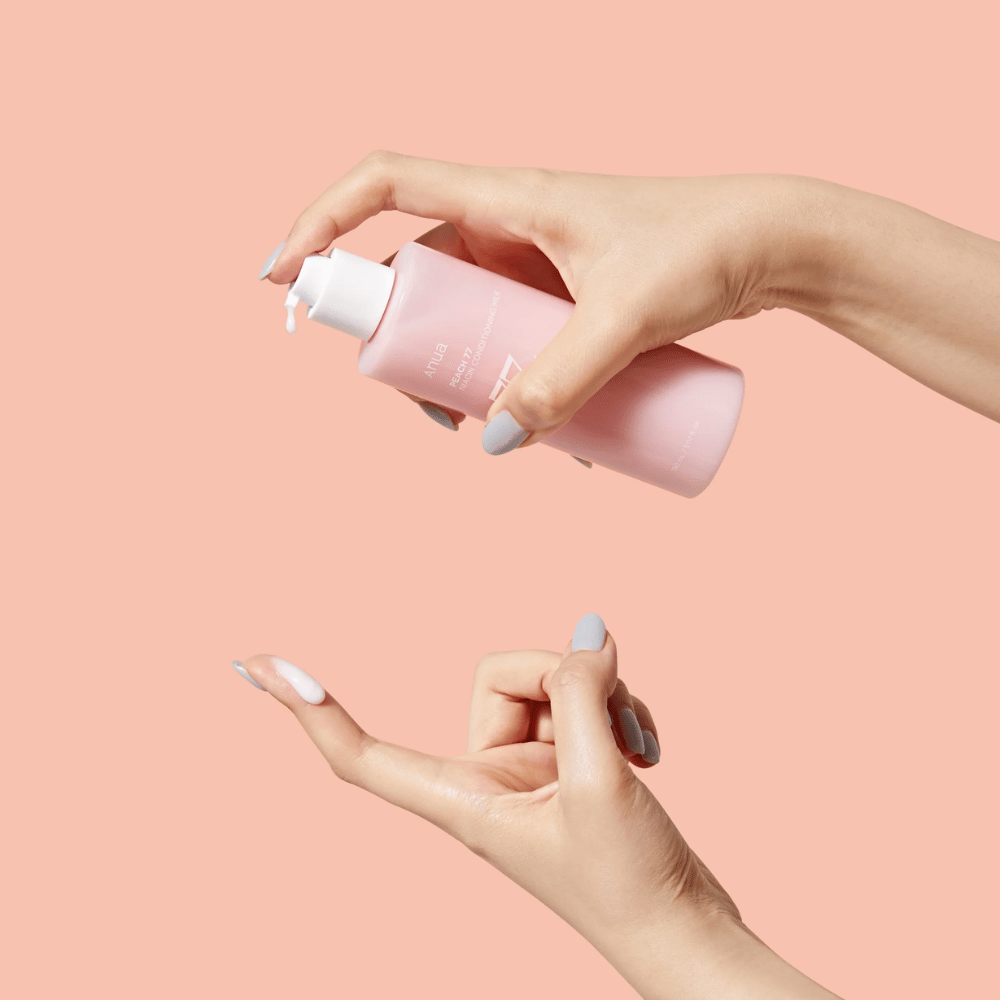 L'image montre les mains de quelqu'un tenant une bouteille avec ANUA Peach 77% Niacin Conditioning Milk. Ce produit de soin de la peau semble avoir une consistance laiteuse et est conçu pour nourrir et améliorer l'état de la peau.