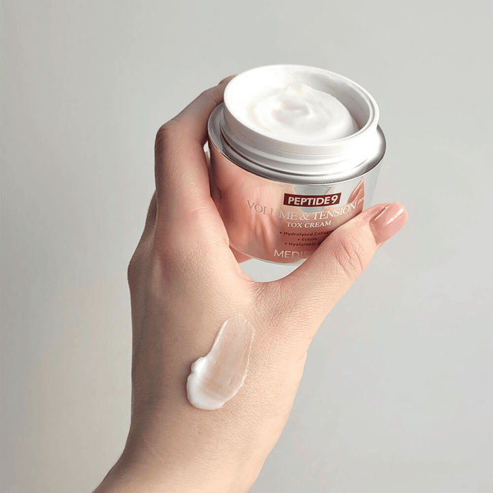 Main tenant MEDI-PEEL Peptide 9 Volume & Tension Tox Cream avec une noisette de crème sur le dos de la main. La formule soyeuse hydrate et donne à la peau fermeté et élasticité.