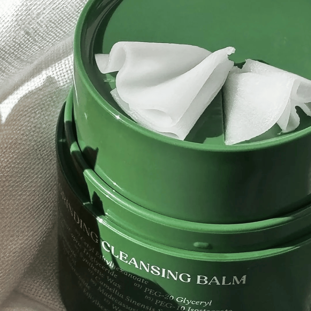 L'image montre un pot vert avec "Cleansing Balm", qui est ouvert pour révéler la spatule incluse et le contenu doux. La consistance crémeuse reflète la lumière, ce qui donne une sensation de luxe et d'efficacité dans le nettoyage.