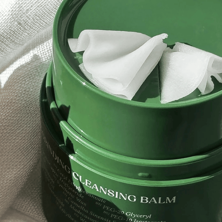 L'image montre un pot vert avec "Cleansing Balm", qui est ouvert pour révéler la spatule incluse et le contenu doux. La consistance crémeuse reflète la lumière, ce qui donne une sensation de luxe et d'efficacité dans le nettoyage.