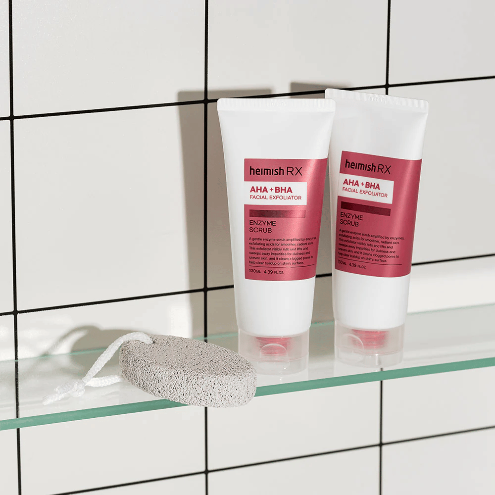 Deux tubes Heimish RX AHA + BHA Facial Exfoliator placés sur une étagère en verre dans une salle de bain avec un mur de carrelage blanc. À côté des produits se trouve une pierre ponce avec une ficelle blanche. Une image de soin de la peau élégante et minimaliste.