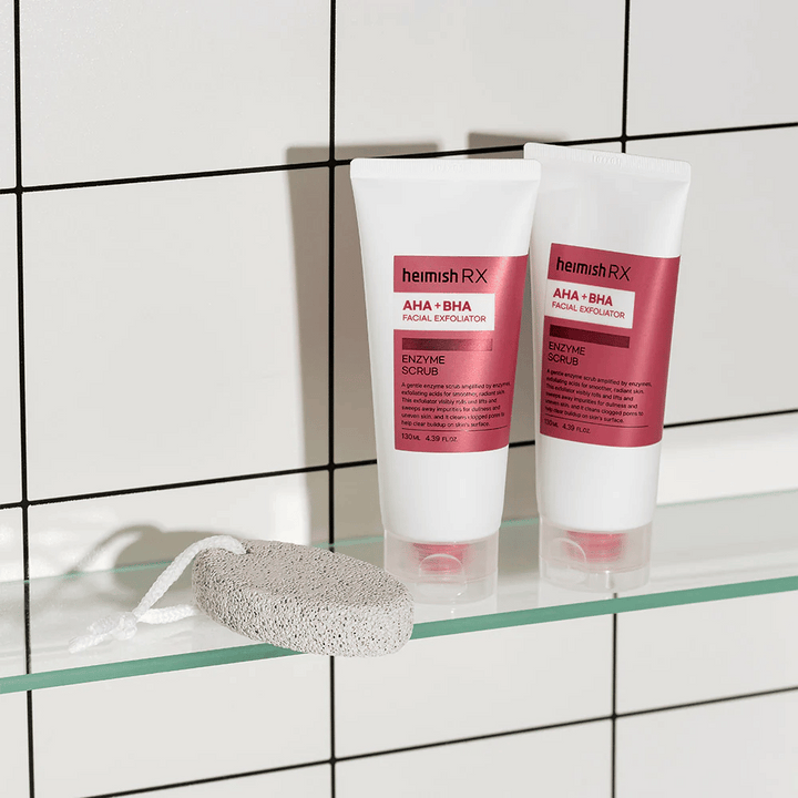 Deux tubes Heimish RX AHA + BHA Facial Exfoliator placés sur une étagère en verre dans une salle de bain avec un mur de carrelage blanc. À côté des produits se trouve une pierre ponce avec une ficelle blanche. Une image de soin de la peau élégante et minimaliste.