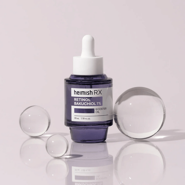 Heimish RX Retinol Bakuchiol Booster Oil 1% dans une élégante bouteille en verre violet avec pipette. L'huile est placée sur une surface réfléchissante avec des billes de verre autour, donnant une sensation luxueuse et moderne.