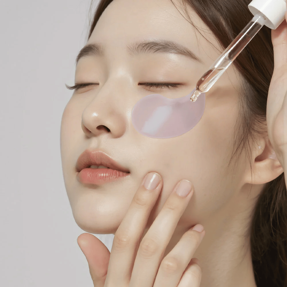 Une femme avec un patch pour les yeux en hydrogel violet applique du sérum sous l'œil avec une pipette. Le produit hydrate et soigne la peau sensible autour des yeux.