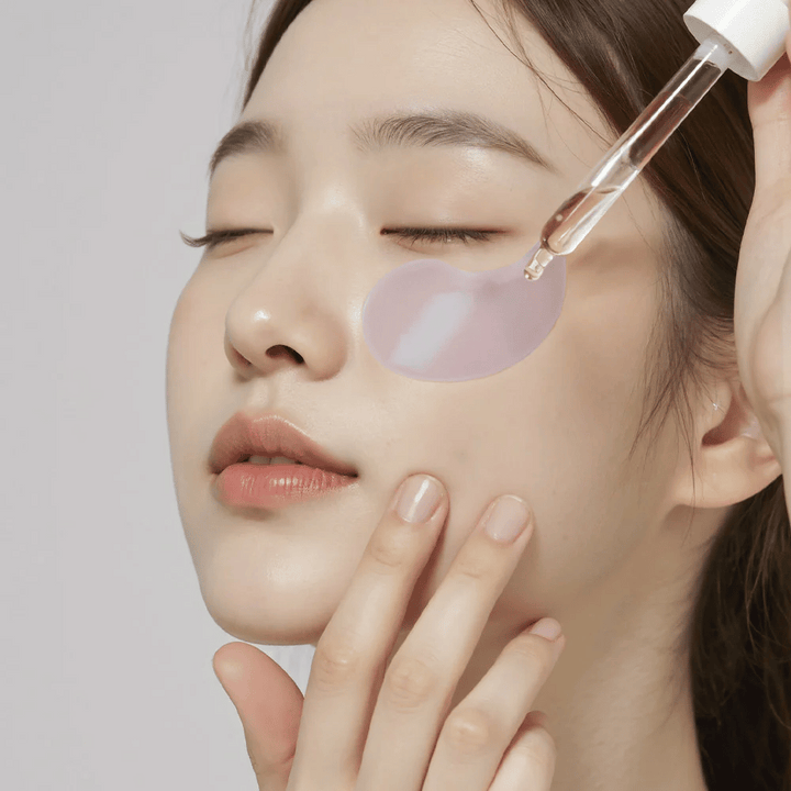 Une femme avec un patch pour les yeux en hydrogel violet applique du sérum sous l'œil avec une pipette. Le produit hydrate et soigne la peau sensible autour des yeux.