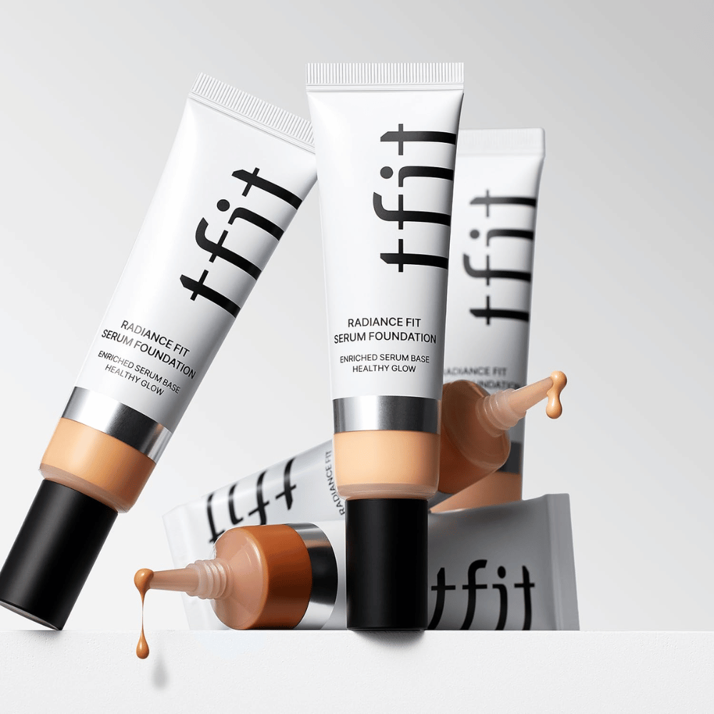Plusieurs tubes de TFIT Radiance Fit Serum Foundation de différentes nuances, dont un avec foundation qui en coule, sur fond gris clair.