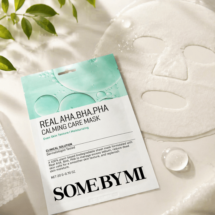 SOME BY MI Real AHA.BHA.PHA Calming Care Mask est présentée avec le masque en tissu déplié. Autour du produit, on voit des gouttes d’hydratation et des feuilles vertes qui renforcent la sensation d’hydratation, de soin apaisant et d’exfoliation douce. L’image met en avant l’accent du masque sur une texture de peau plus uniforme et un meilleur équilibre hydrique.
