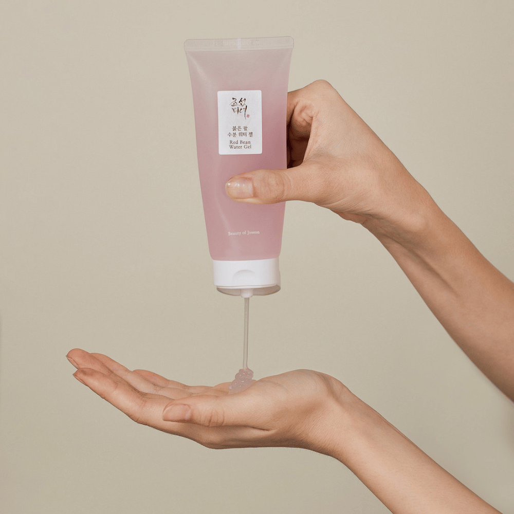 L'image montre une personne tenant un tube de "Red Bean Water Gel" de Beauty of Joseon et pressant un gel transparent dans sa main. Le produit semble être destiné aux soins de la peau et a un emballage attrayant, de couleur rose doux.