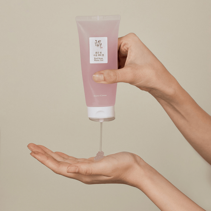 L'image montre une personne tenant un tube de "Red Bean Water Gel" de Beauty of Joseon et pressant un gel transparent dans sa main. Le produit semble être destiné aux soins de la peau et a un emballage attrayant, de couleur rose doux.