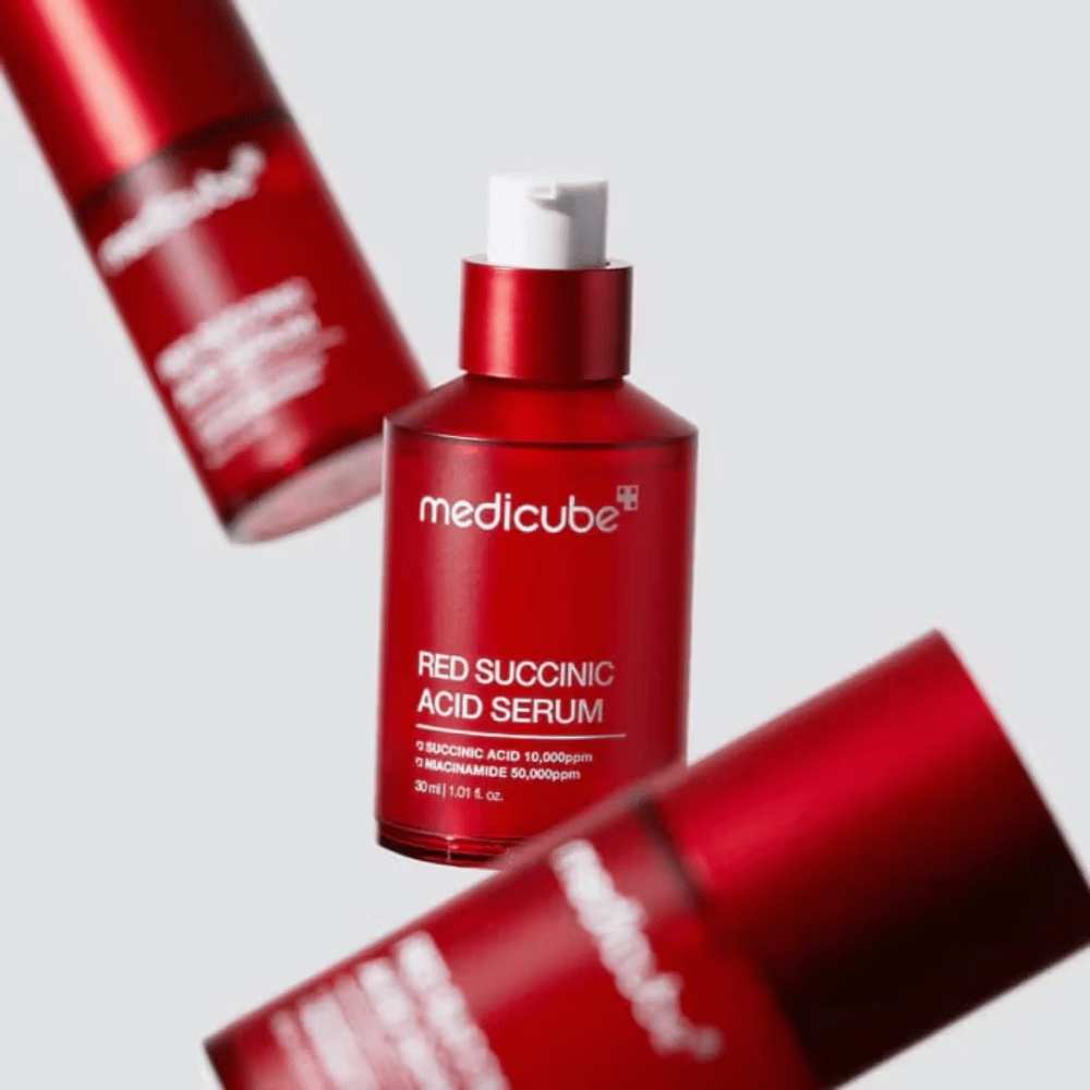 Medicube Sérum à l'acide succinique rouge dans un flacon pompe rouge, entouré de plusieurs flacons identiques sur un fond clair. Le produit contient de l'acide succinique et de la niacinamide pour traiter l'acné et les taches pigmentaires.