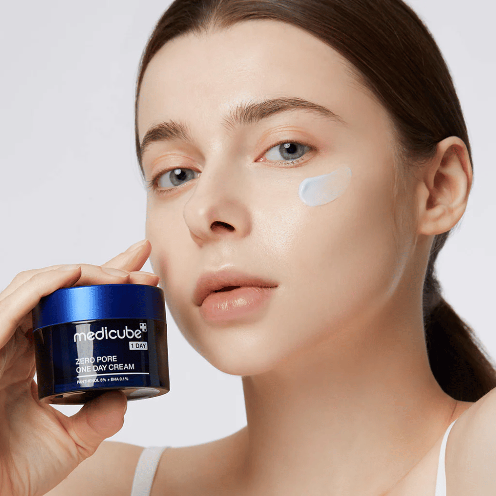 Femme avec un look naturel tient Medicube Zero Pore One Day Cream. Une petite quantité de crème est appliquée sur sa joue. Le produit est conçu pour améliorer la texture de la peau et minimiser la taille des pores, avec une formule légère et hydratante.