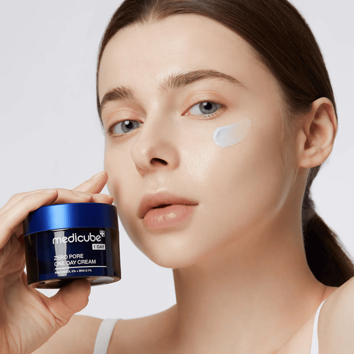 Femme avec un look naturel tient Medicube Zero Pore One Day Cream. Une petite quantité de crème est appliquée sur sa joue. Le produit est conçu pour améliorer la texture de la peau et minimiser la taille des pores, avec une formule légère et hydratante.