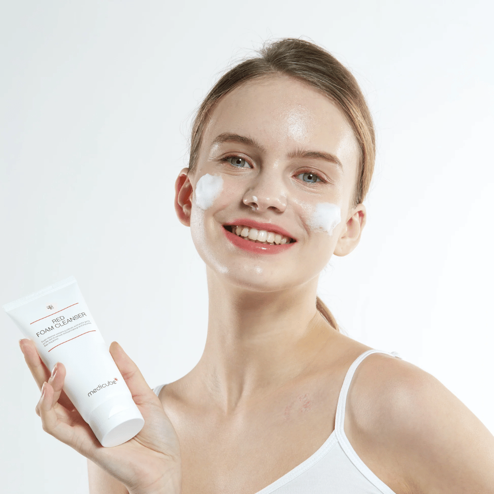 Jeune femme avec de la mousse nettoyante sur le visage tenant un tube de Medicube Red Foam Cleanser et souriant à la caméra. Elle porte un débardeur blanc et le fond est clair et simple.