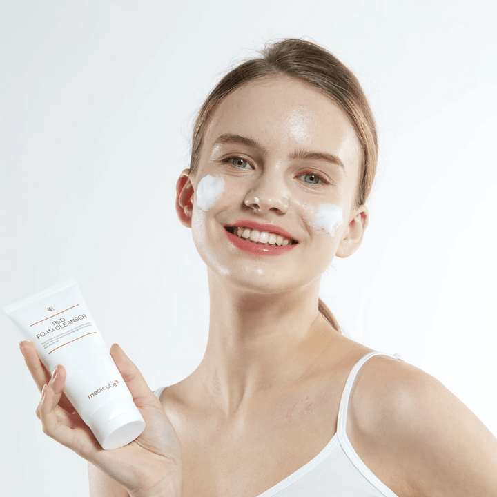 Jeune femme avec de la mousse nettoyante sur le visage tenant un tube de Medicube Red Foam Cleanser et souriant à la caméra. Elle porte un débardeur blanc et le fond est clair et simple.