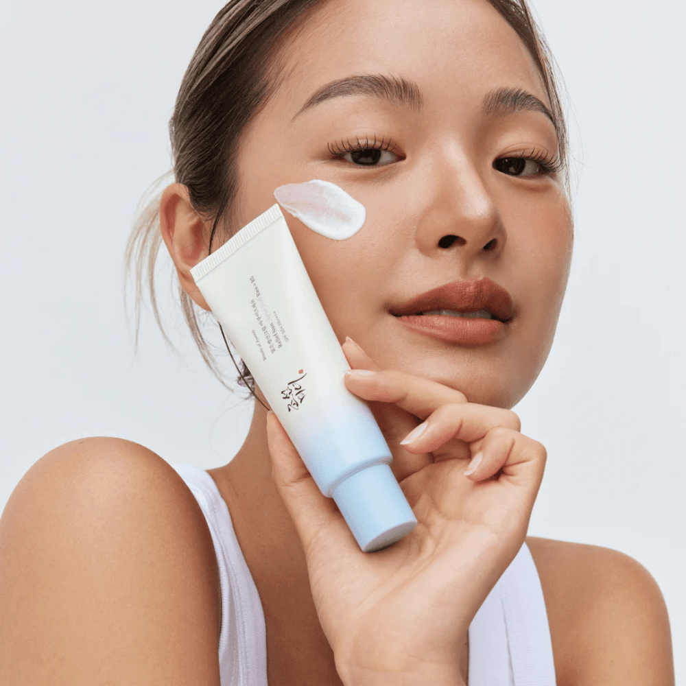 Une femme tient Beauty of Joseon Relief Sun : crème solaire Aqua-Fresh Rice + B5 et montre sa consistance crémeuse sur la joue. Un produit solaire léger qui hydrate et protège la peau avec SPF 50+ PA++++.