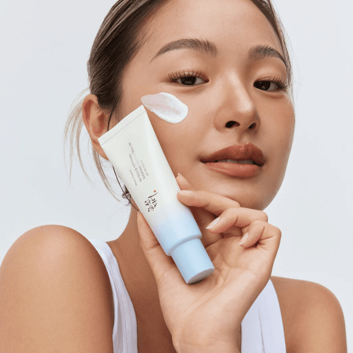 Une femme tient Beauty of Joseon Relief Sun : crème solaire Aqua-Fresh Rice + B5 et montre sa consistance crémeuse sur la joue. Un produit solaire léger qui hydrate et protège la peau avec SPF 50+ PA++++.