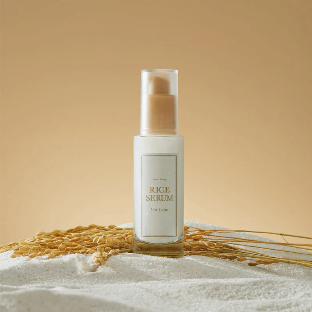 Bouteille avec "I'm from" Rice Serum, entourée de grains de riz et d'épis de riz sur un fond chaud.