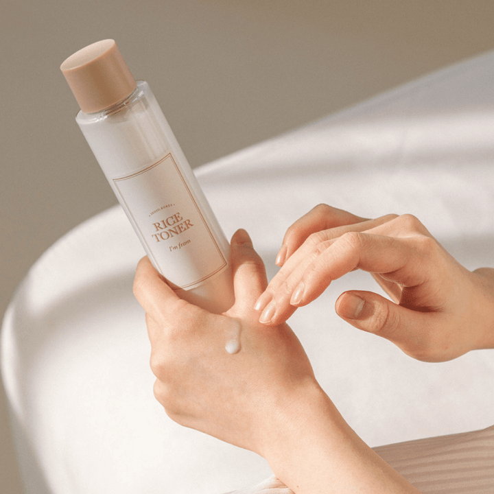 Des mains tiennent une bouteille de lotion tonique au riz marquée "I'm from". Une goutte du produit est appliquée sur une main tandis que l'autre main se prépare à l'appliquer. La scène est doucement éclairée avec une teinte chaude.