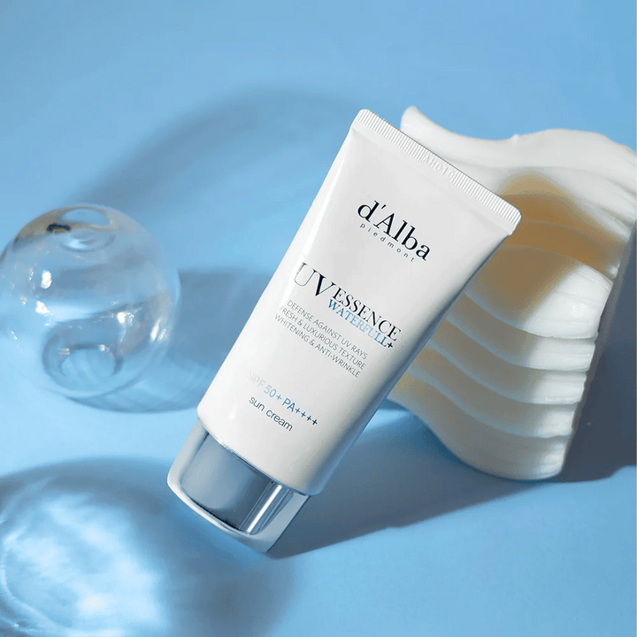 d'Alba UV Essence Waterfull+ Sun Cream avec SPF 50+ PA++++ est posé sur un fond bleu avec des reflets lumineux et des éléments décoratifs – met en avant les propriétés fraîches et hydratantes de la crème solaire.