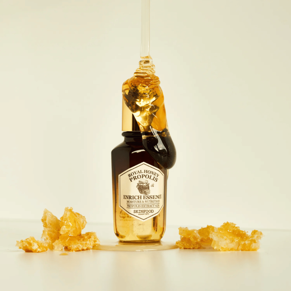 Image d'une bouteille en verre avec Royal Honey Propolis Enrich Essence de Skinfood. Une grande goutte de miel doré coule du bouchon doré de la bouteille. Autour de la bouteille, de petits morceaux de rayon de miel sont dispersés sur une surface claire. L'essence est commercialisée avec un accent sur l'hydratation et la nutrition, avec 63% d'extrait de propolis.