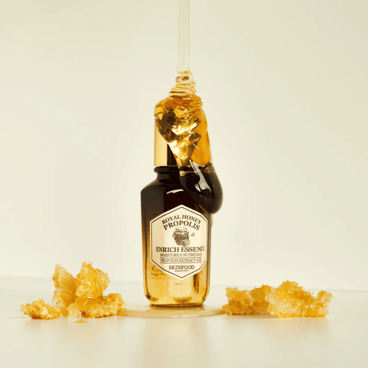 Image d'une bouteille en verre avec Royal Honey Propolis Enrich Essence de Skinfood. Une grande goutte de miel doré coule du bouchon doré de la bouteille. Autour de la bouteille, de petits morceaux de rayon de miel sont dispersés sur une surface claire. L'essence est commercialisée avec un accent sur l'hydratation et la nutrition, avec 63% d'extrait de propolis.