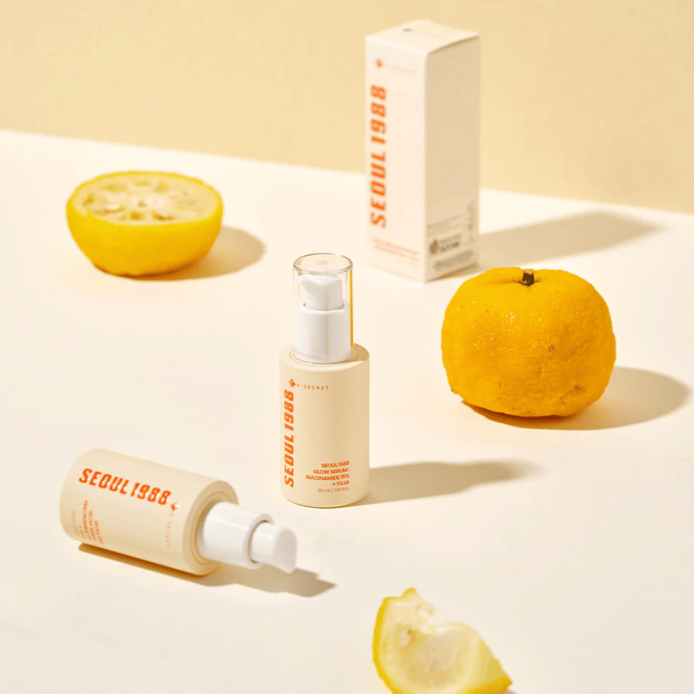Plusieurs bouteilles et emballages de Séoul 1988 Glow Serum arrangés avec des fruits yuja entiers et coupés en deux en arrière-plan.