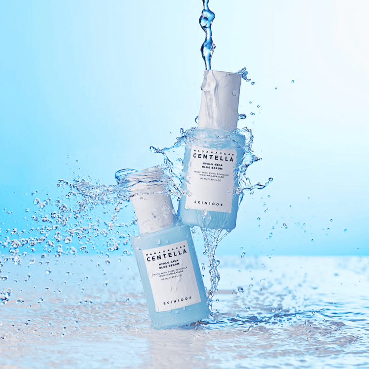 Deux bouteilles de SKIN1004 Madagascar Centella Hyalu-Cica Blue Serum sont frappées par une cascade d'eau sur un fond bleu clair.