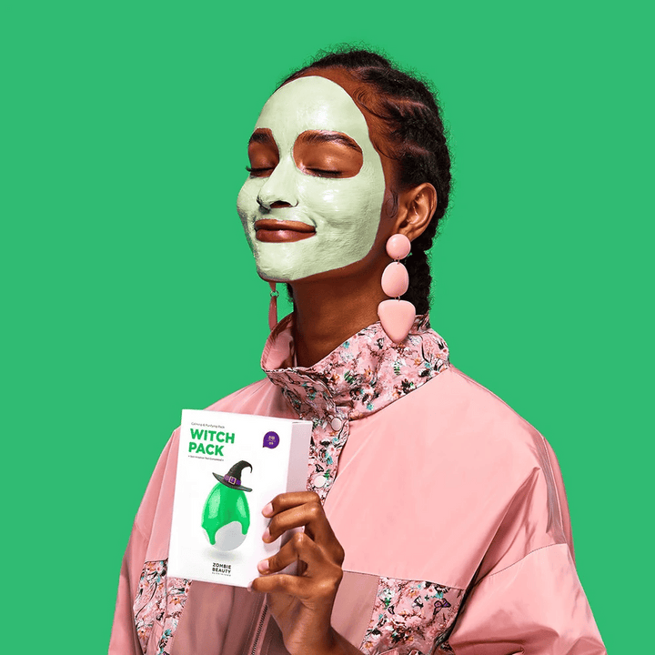 Une femme avec un masque facial vert de Zombie Beauty Witch Pack sourit et tient l'emballage dans sa main. Elle porte une veste rose avec un motif floral et de grandes boucles d'oreilles roses. L'arrière-plan est vert et correspond à la couleur du masque facial.