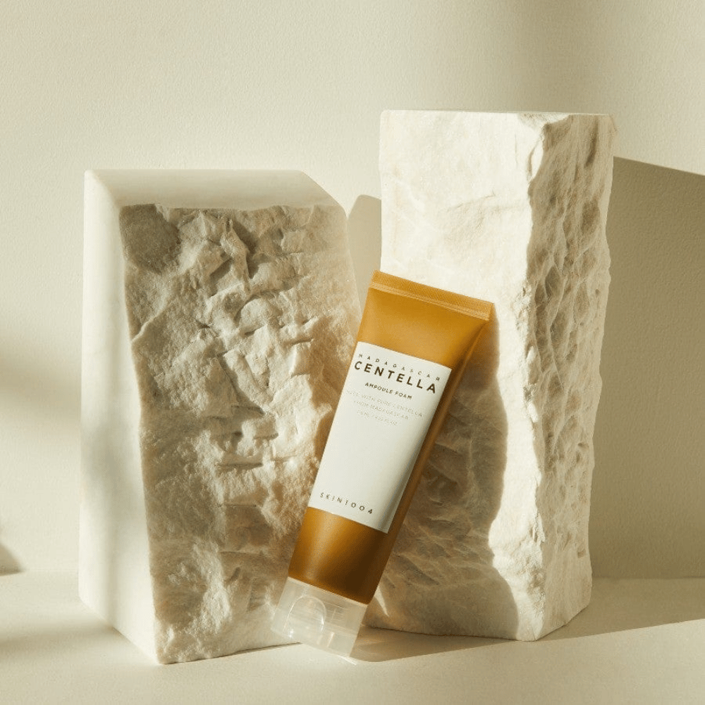 SKIN1004 Madagascar Centella Ampoule Foam debout contre des blocs de pierre clairs à la lumière naturelle du soleil, avec leur design tubulaire brun et blanc et une élégante minimalisme.