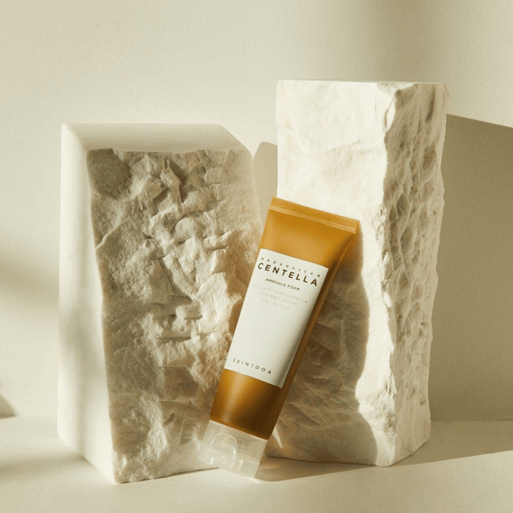 SKIN1004 Madagascar Centella Ampoule Foam debout contre des blocs de pierre clairs à la lumière naturelle du soleil, avec leur design tubulaire brun et blanc et une élégante minimalisme.