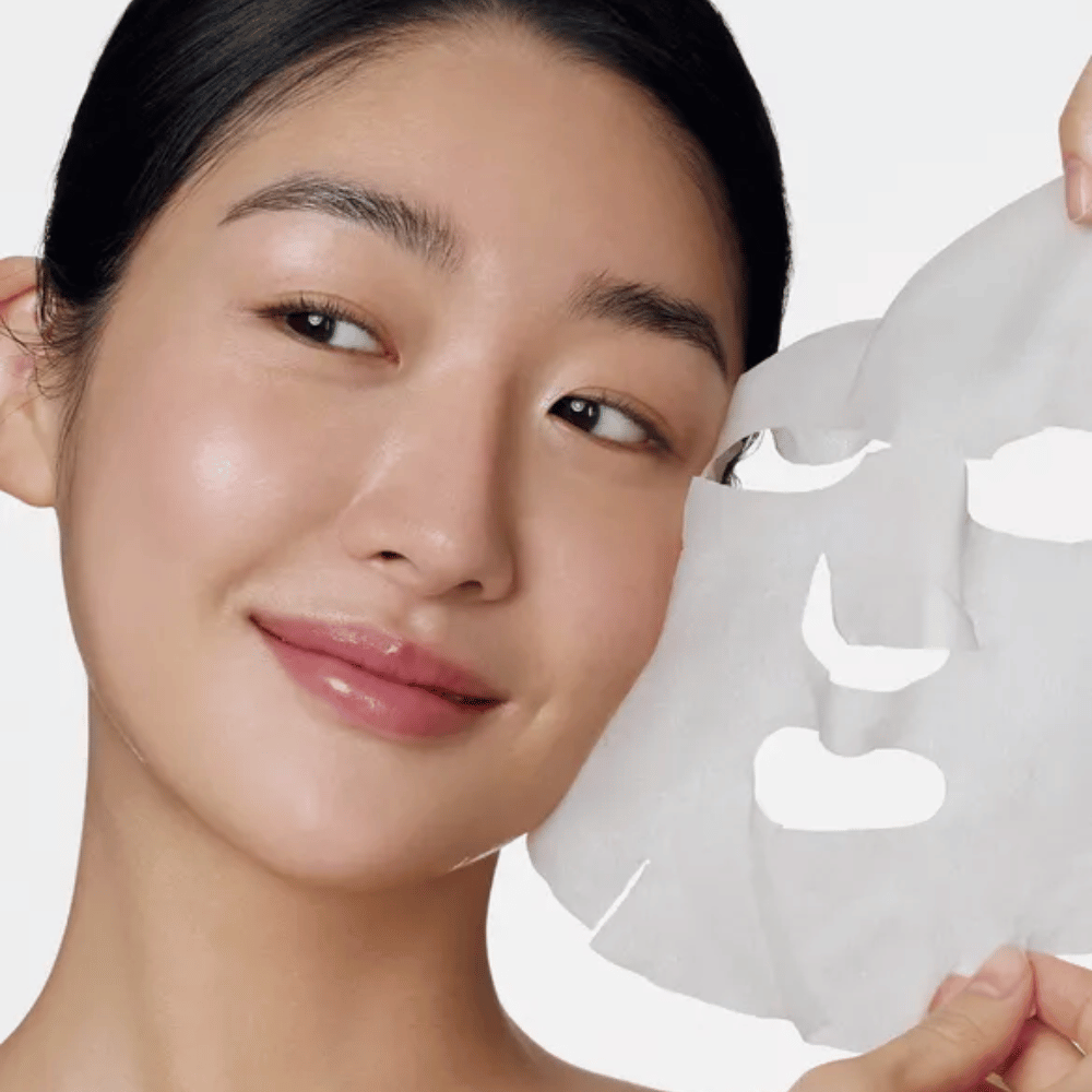 L'image montre une personne utilisant un masque facial, spécifiquement un "Retinol Intense Daily Mask" de Some By Mi. Le masque est conçu pour une utilisation quotidienne et contient du rétinol pour améliorer la texture de la peau, réduire les ridules et donner un teint plus uniforme.
