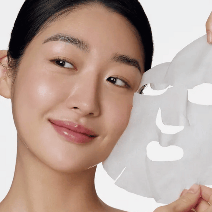 L'image montre une personne utilisant un masque facial, spécifiquement un "Retinol Intense Daily Mask" de Some By Mi. Le masque est conçu pour une utilisation quotidienne et contient du rétinol pour améliorer la texture de la peau, réduire les ridules et donner un teint plus uniforme.