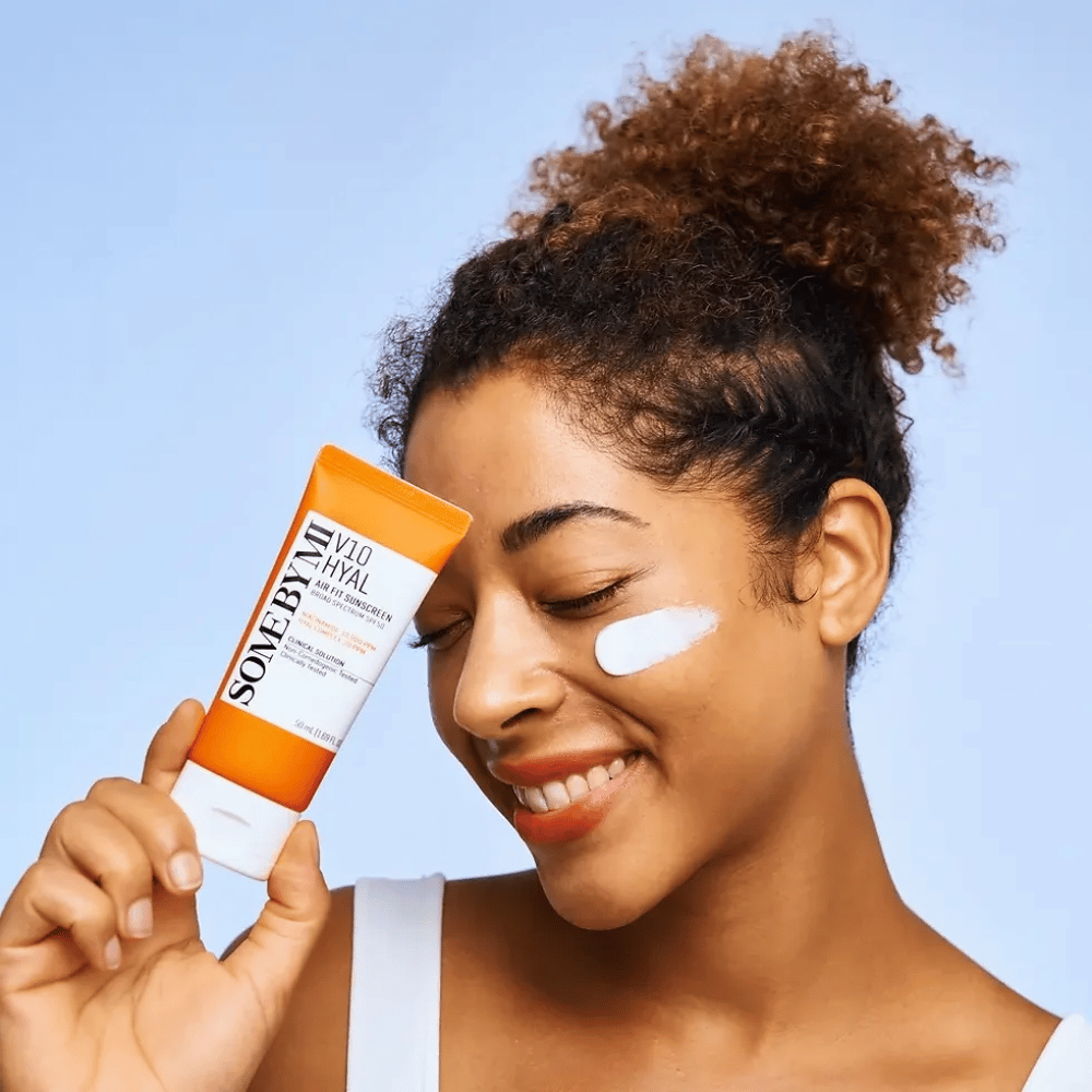 Une femme tient un tube de SOME BY MI V10 Hyal Air Fit Sunscreen et a une tache blanche de crème solaire sur la joue. Elle sourit joyeusement et montre clairement le produit, mettant en avant son utilisation et ses avantages pour le soin de la peau et la protection contre le soleil.