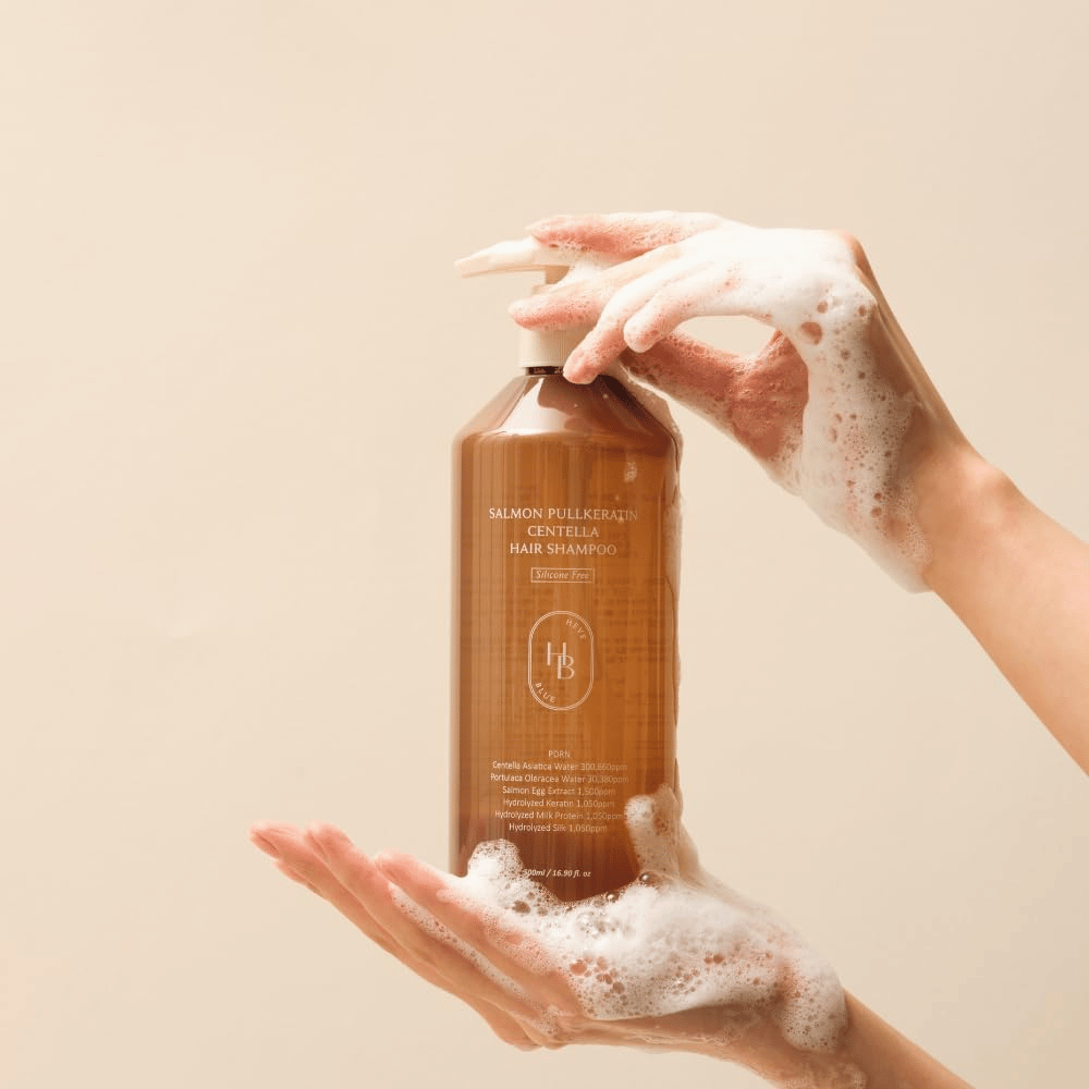 Heveblue Salmon Pullkeratin Centella Hair Shampoo dans un flacon brun entouré de mousse. Le produit est tenu entre deux mains et met en avant une expérience de soin capillaire nettoyante et nourrissante avec une mousse abondante.