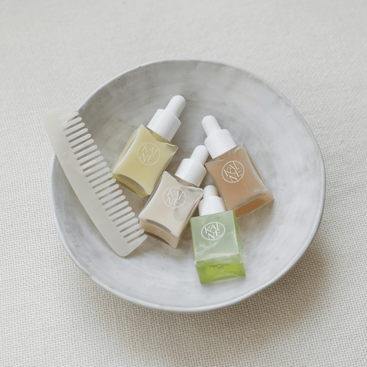 Quatre petits flacons de sérum de KAINÉ Serum Discovery Kit sont dans un bol blanc à côté d'un peigne, avec un arrière-plan naturel et apaisant en ton beige.