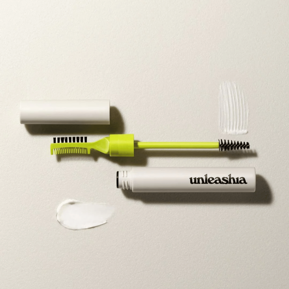 Un ensemble de sculpture pour sourcils avec un peigne vert, une brosse noire et un tube blanc marqué "unleashia" sur un fond clair avec une masse de produit transparent.