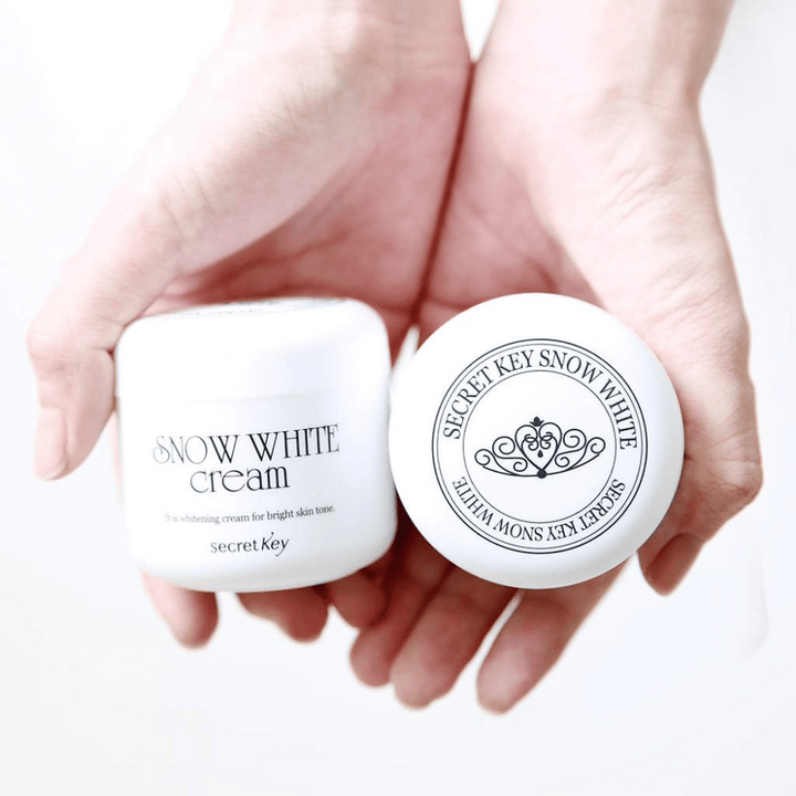 Deux pots de Secret Key Snow White Cream tenus dans les mains, un montrant le couvercle et un montrant l'avant.