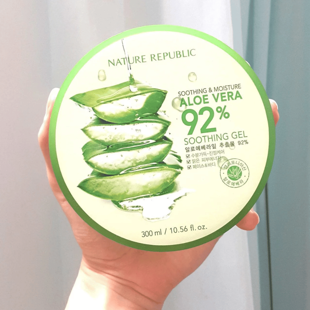 Une main tient une boîte ronde avec Nature Republic Aloe Vera 92% Gel Apaisant. La boîte est verte avec une image d'aloe vera et un texte décrivant ses propriétés apaisantes et hydratantes. Le contenu est de 300 ml.