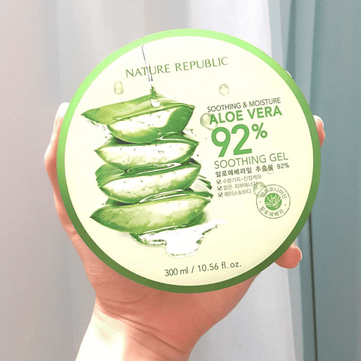 Une main tient une boîte ronde avec Nature Republic Aloe Vera 92% Gel Apaisant. La boîte est verte avec une image d'aloe vera et un texte décrivant ses propriétés apaisantes et hydratantes. Le contenu est de 300 ml.