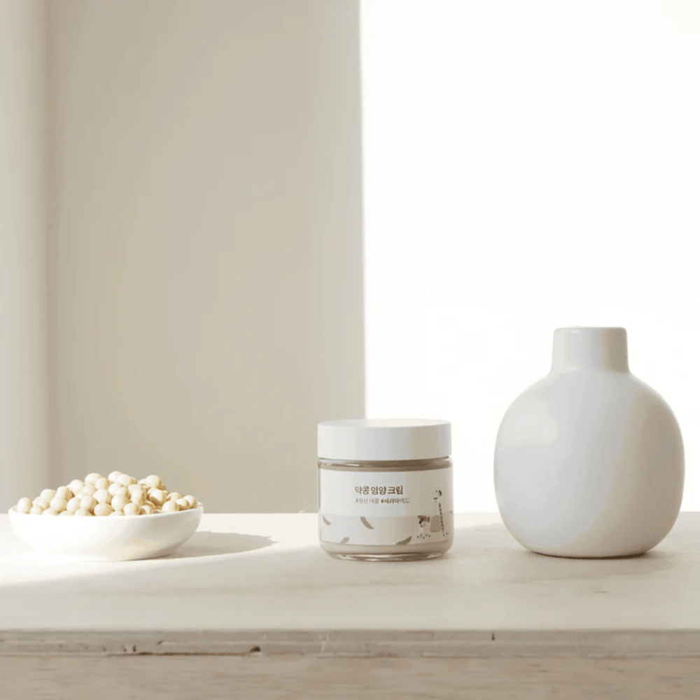 Mixsoon Soybean Nourishing Cream se tient sur une table en bois à côté d'un vase et de fèves de soja. Une crème minimaliste et hydratante avec des ingrédients naturels.