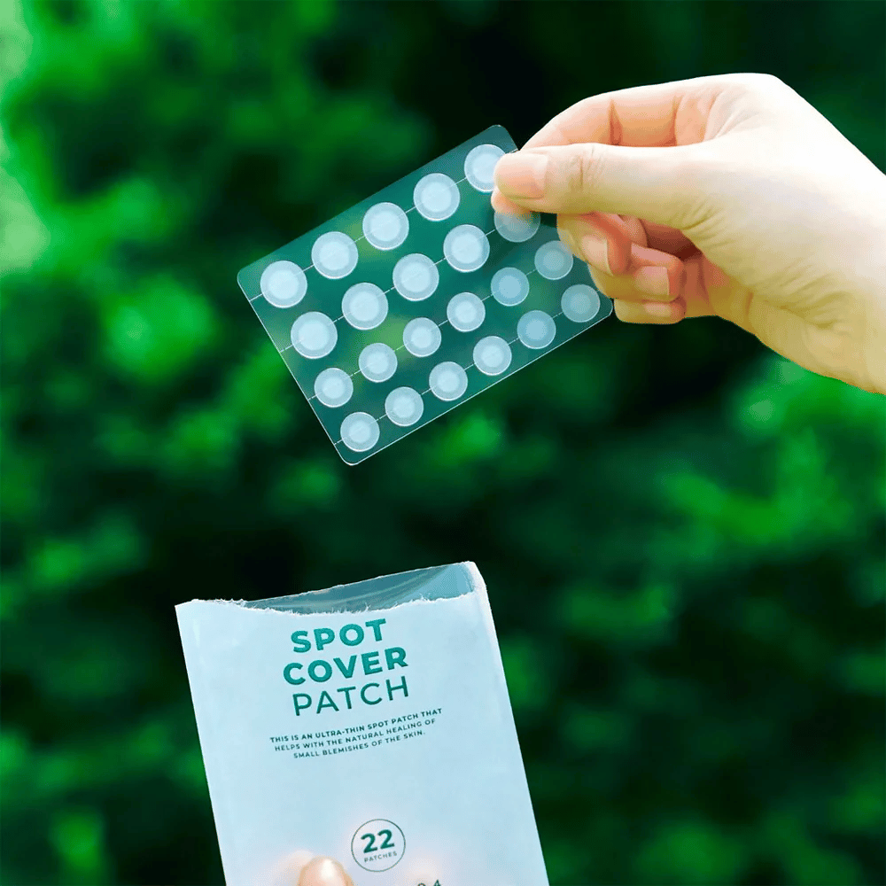 Une main tient une feuille de Spot Cover Patches, qui sont transparents et arrondis. En arrière-plan, on voit un environnement vert flou. L'emballage montre une partie de l'emballage avec le texte "SPOT COVER PATCH" et des informations indiquant que le produit contient 22 pansements qui aident à guérir de petites zones de peau.