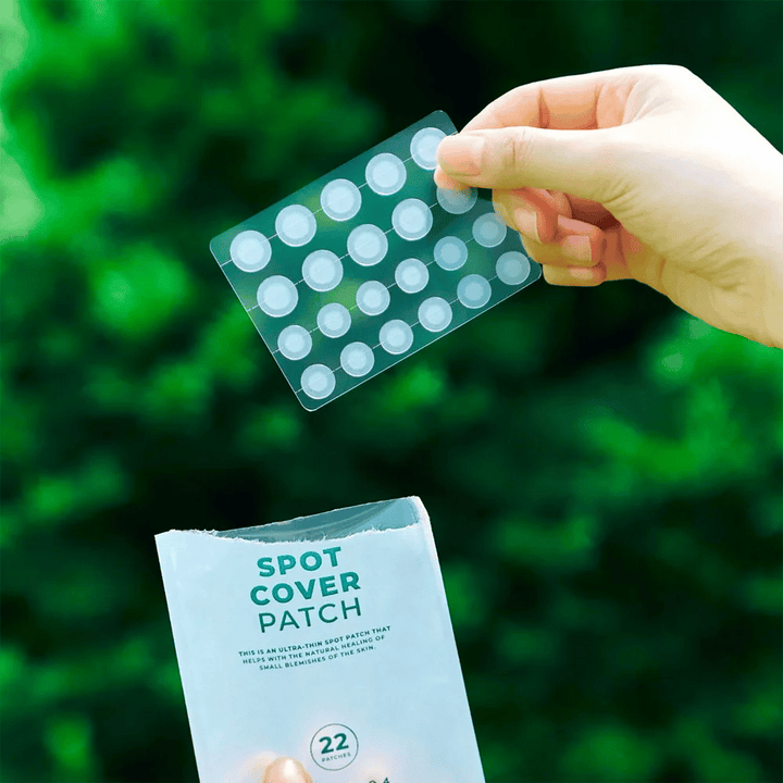 Une main tient une feuille de Spot Cover Patches, qui sont transparents et arrondis. En arrière-plan, on voit un environnement vert flou. L'emballage montre une partie de l'emballage avec le texte "SPOT COVER PATCH" et des informations indiquant que le produit contient 22 pansements qui aident à guérir de petites zones de peau.