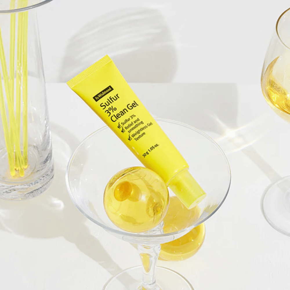 By Wishtrend Sulfur 3% Clean Gel dans un tube jaune placé dans un verre à cocktail avec une sphère jaune, entouré de verres élégants sur un fond blanc.