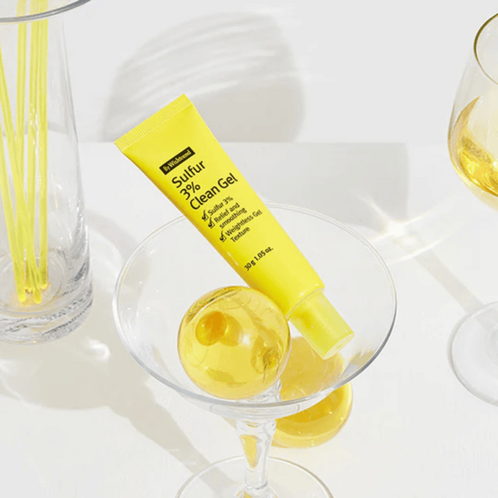 By Wishtrend Sulfur 3% Clean Gel dans un tube jaune placé dans un verre à cocktail avec une sphère jaune, entouré de verres élégants sur un fond blanc.