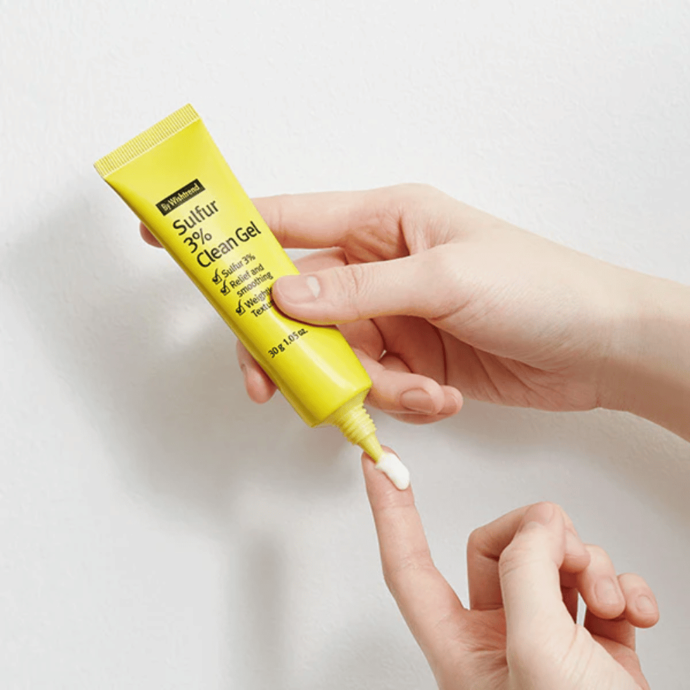 Gros plan sur des mains appliquant By Wishtrend Sulfur 3% Clean Gel, un tube jaune avec du gel blanc, sur un fond blanc.