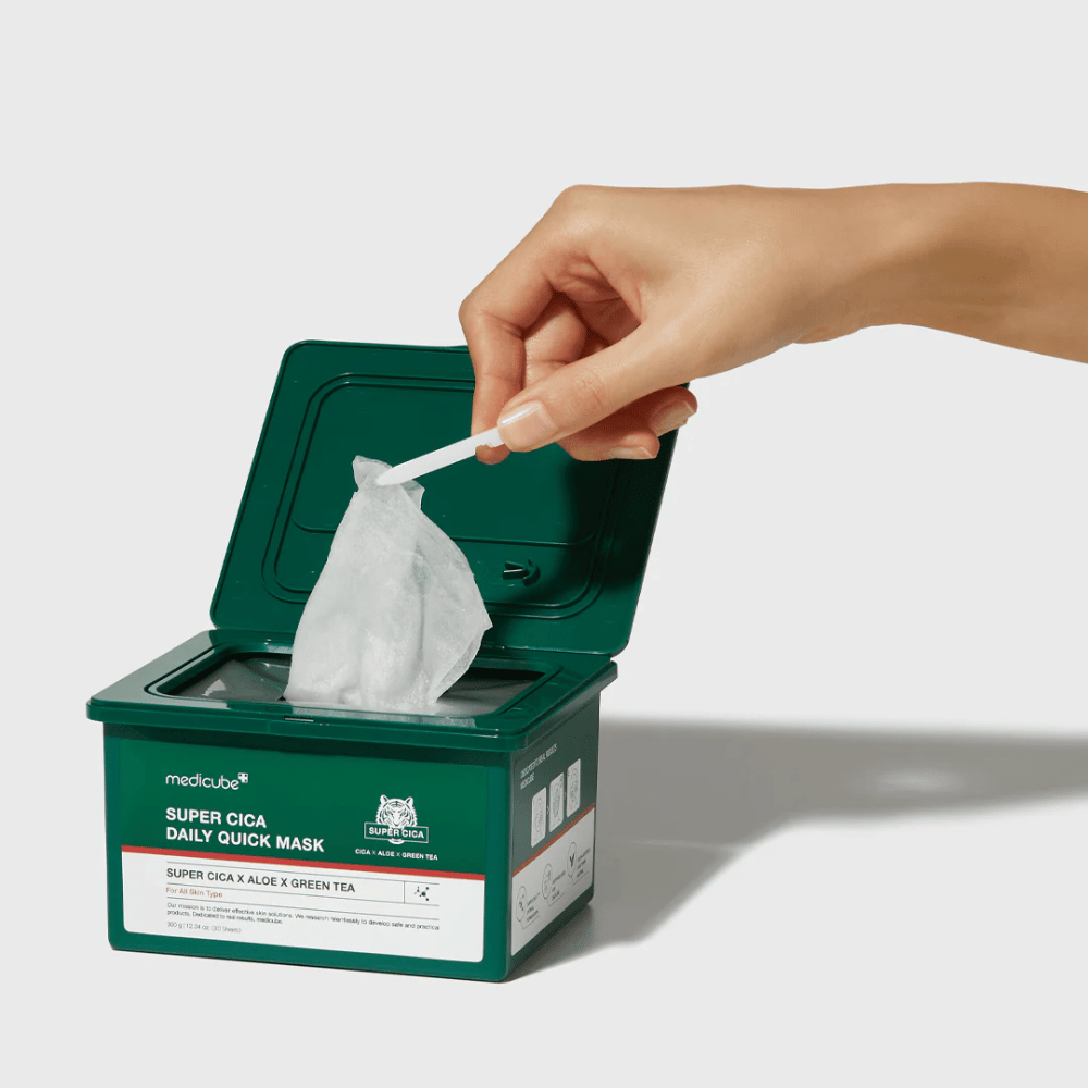 Contenant vert avec couvercle ouvert montrant medicube Super Cica Daily Quick Mask. Une main retire un masque en feuille avec une pince incluse. Le produit contient Cica, aloe et thé vert, idéal pour une hydratation rapide et un effet apaisant.