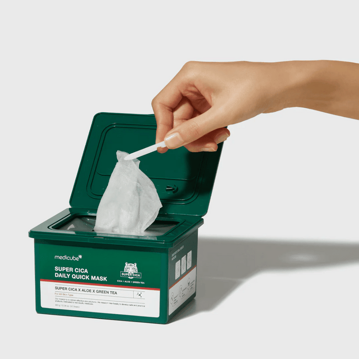 Contenant vert avec couvercle ouvert montrant medicube Super Cica Daily Quick Mask. Une main retire un masque en feuille avec une pince incluse. Le produit contient Cica, aloe et thé vert, idéal pour une hydratation rapide et un effet apaisant.